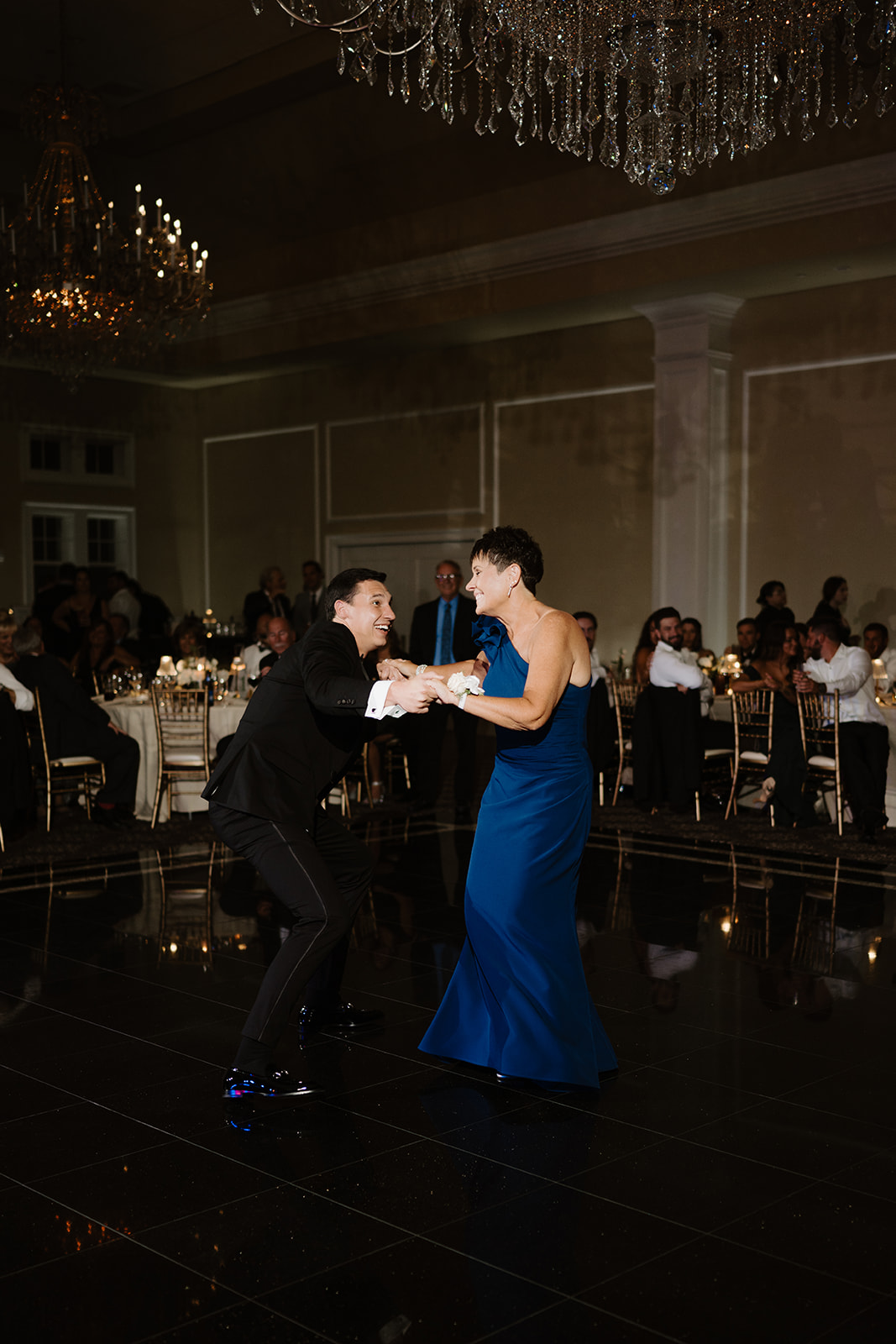 Groom and his mother in a blue gown sharing a fun, lively dance on the reflective ballroom floor.